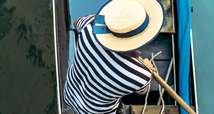 Top-down view of a gondolier in striped shirt and straw hat rowing a gondola through green canal water.