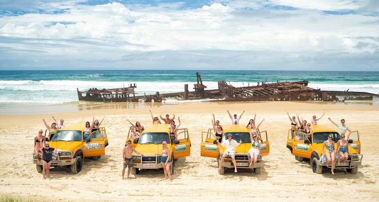 Group of people posing with 4WD vehicles on a beach near a shipwreck.