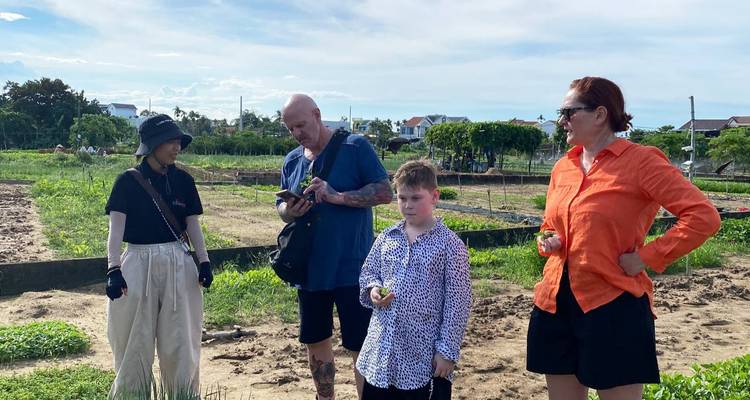 Visitantes y guía local de pie en un huerto recogiendo productos bajo un cielo azul