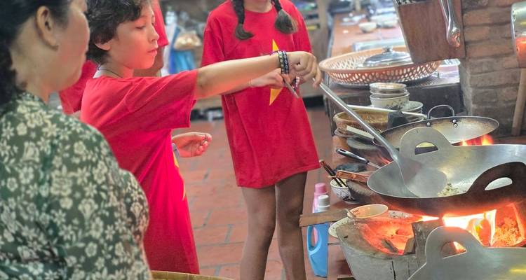 Enfants vêtus de chemises rouges cuisinant dans des woks traditionnels alimentés au feu de bois lors d'une classe pratique.