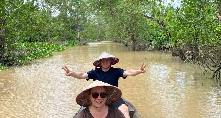 Les touristes portant des chapeaux coniques posant avec des signes de paix dans un bateau étroit sur un canal boueux du Mékong.