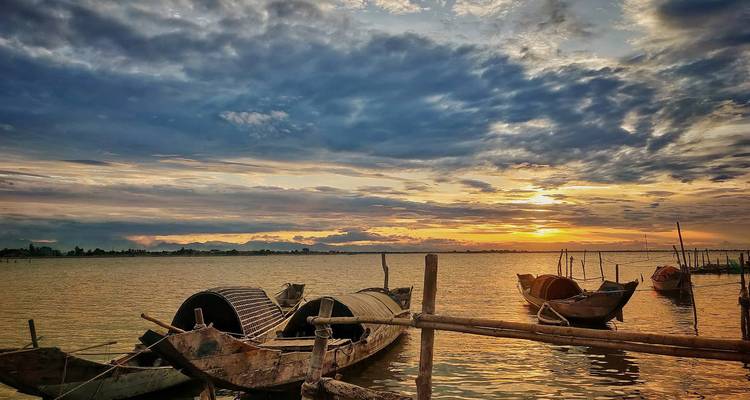 Des bateaux de pêche en bois reposent près de poteaux en bambou sur des eaux calmes tandis que des nuages spectaculaires s'illuminent au coucher du soleil.