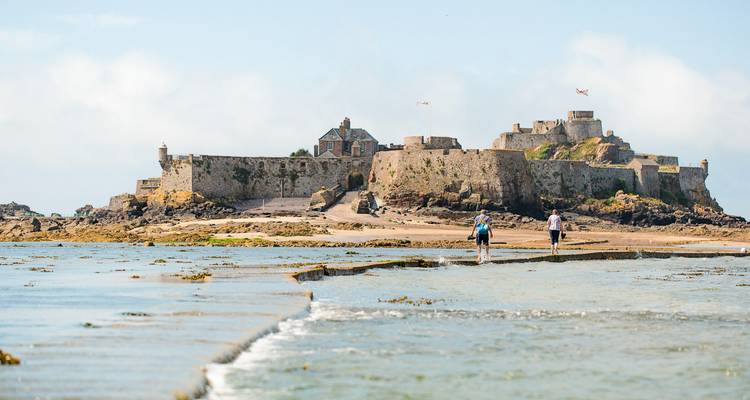 Two walkers cross a tidal causeway leading to a stone fort on a small island.