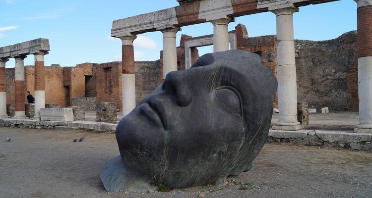 Grande tête de statue gisant au sol devant des colonnes antiques.