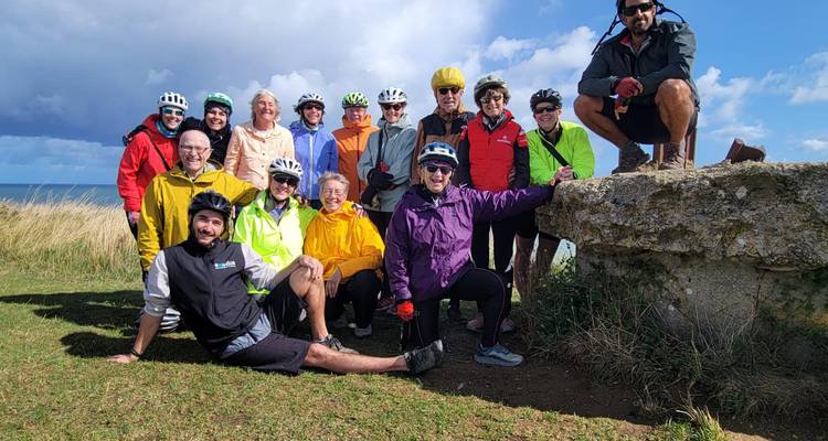 Grote vrolijke fietsgroep die poseert op een grasparkachtige klif met helmen, fietsen en een zeegezicht op de achtergrond.