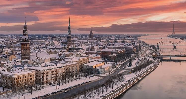 Ciudad vieja de Riga cubierta de nieve y puentes del río bajo un dramático cielo rosado del atardecer.