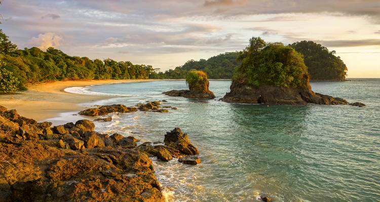 Crique tropicale isolée avec sable doré, verdure de la jungle et îlots rocheux au coucher du soleil.