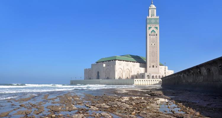 Mosquée au bord de l'océan avec des vagues qui se brisent sur les rochers sous un ciel bleu dégagé.