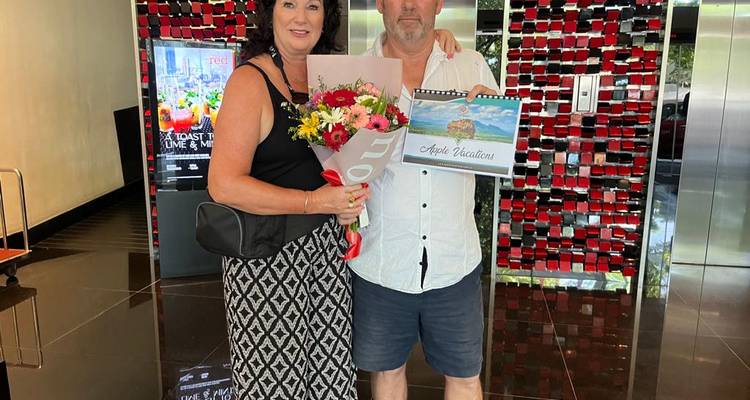 Two people posing with flowers and a sign indoors.