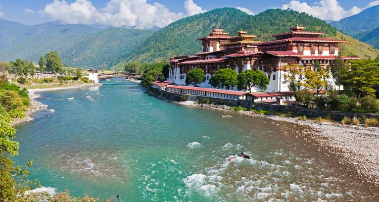 A large traditional Bhutanese building by a river surrounded by mountains.