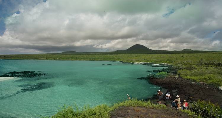Bahía turquesa bordeada por una costa de lava negra donde un pequeño grupo se prepara para hacer snorkel bajo nubes dramáticas.