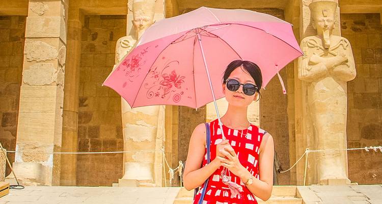 Une touriste en robe rouge tient un parapluie rose devant des statues de gardiens dans un temple égyptien antique.