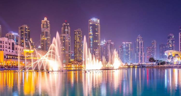 Nächtliche Dubai-Fontänenshow mit illuminierten Wasserstrahlen, die vor einer glänzenden Skyline aufsteigen und sich im See spiegeln.