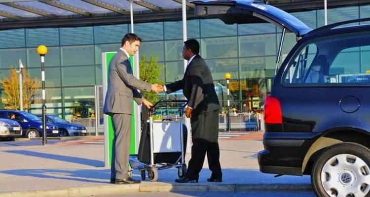 Ein Geschäftsreisender schüttelt die Hand eines Chauffeurs neben einem schwarzen Auto vor einem Flughafenterminal.