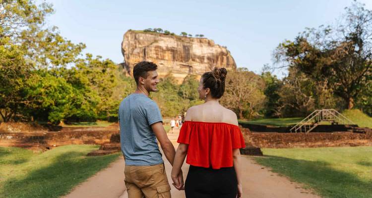 Junges Paar hält Händchen beim Spaziergang in Richtung des ikonischen Sigiriya-Felsens, der sich über üppiges Parkland erhebt.