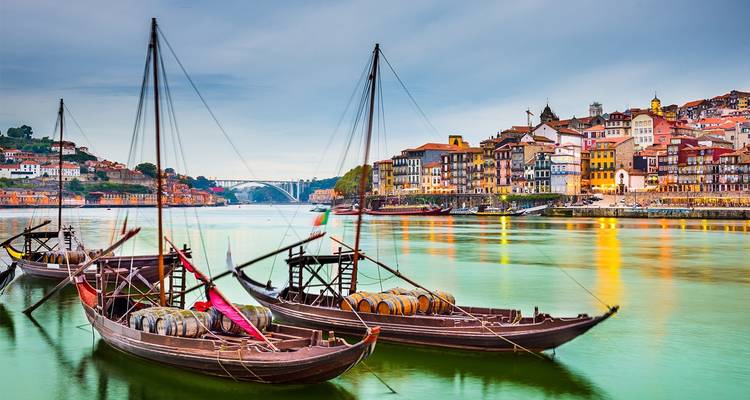 Traditionelle Rabelo-Boote, beladen mit Weinfässern, gleiten bei Einbruch der Dämmerung auf dem Fluss Douro gegenüber der farbenfrohen historischen Uferpromenade von Porto.