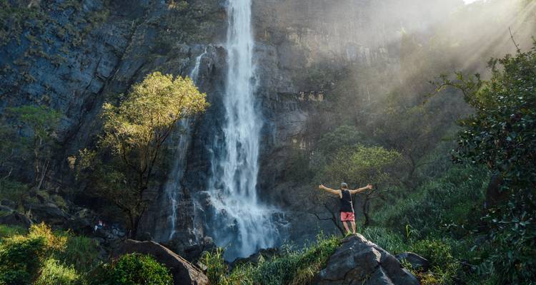 Viajero con los brazos extendidos celebra frente a una alta cascada de la selva que cae por un acantilado rocoso.