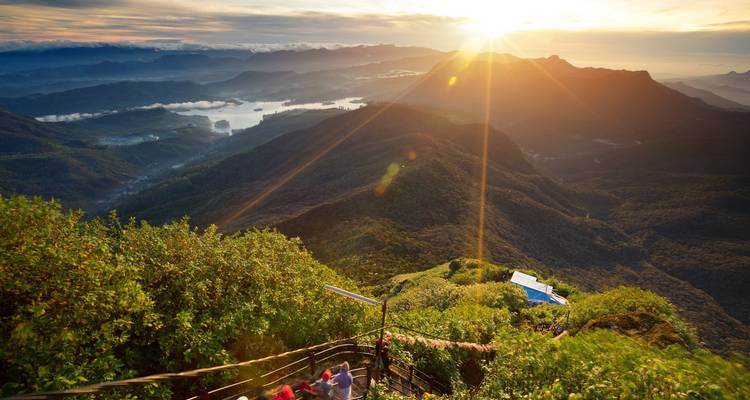 Vista panorámica del amanecer desde la cima de una montaña con rayos de sol sobre valles verdes escalonados y lagos distantes.