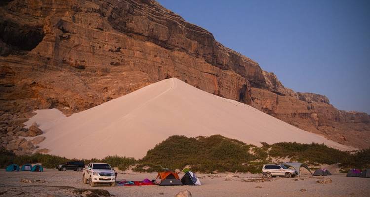 Acampada grupal en la base de una gran duna de arena con autos estacionados cerca.