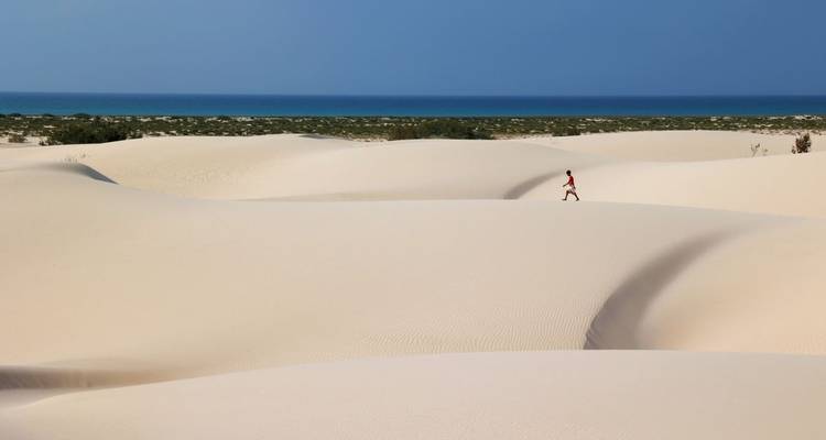 Persona caminando a través de dunas de arena con el océano de fondo.