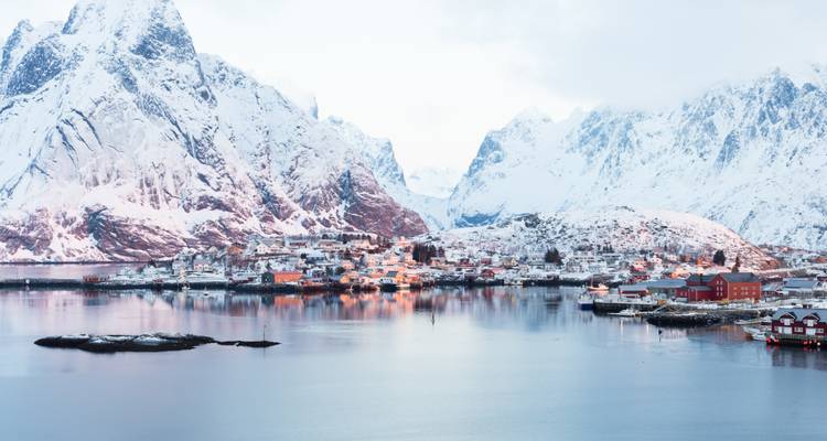 Village enneigé niché entre des pics spectaculaires se reflétant dans le fjord.