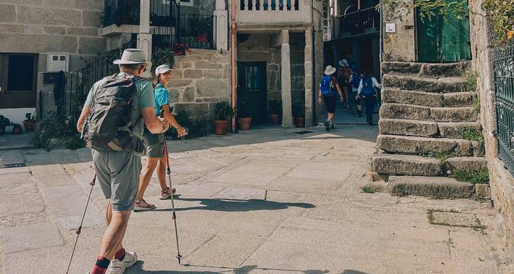 Caminantes mayores usando bastones de trekking pasando por casas de piedra en un callejón del pueblo.