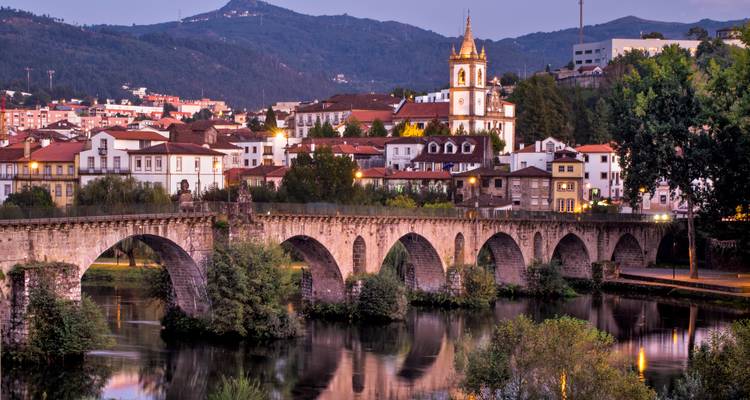 Pont en pierre aux arches historiques traversant une rivière tranquille avec une ville pittoresque et un clocher d'église illuminés au crépuscule.