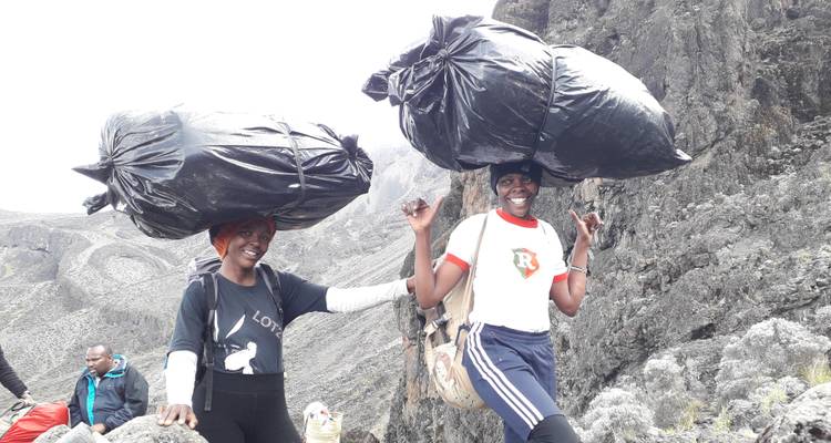 Deux porteuses souriantes transportant de grands ballots noirs sur leur tête en parcourant un terrain montagneux rocheux lors d'une randonnée.