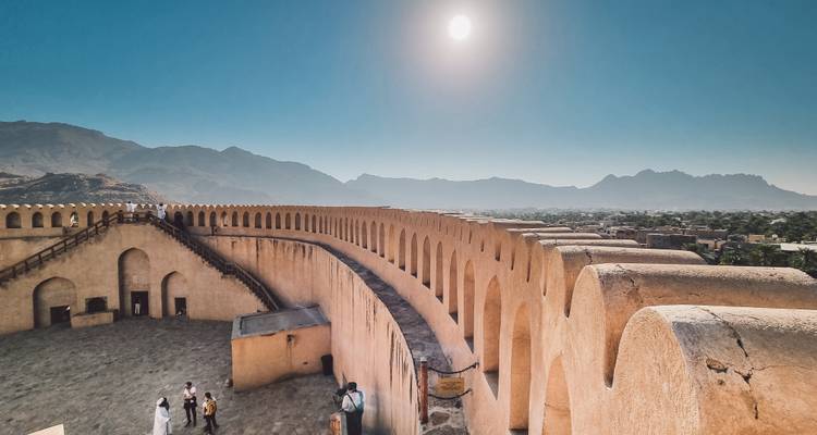 Les visiteurs explorent les remparts du fort de Nizwa sous un soleil intense avec des montagnes brumeuses au loin.