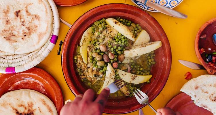 Bovenaanzicht van een kleurrijke Marokkaanse tajine met erwten, aardappelen en olijven op een felgele tafel.
