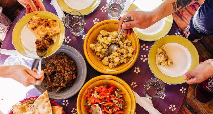 Overhead view of a table set with Moroccan dishes, flatbreads and colorful salads as hands serve food.