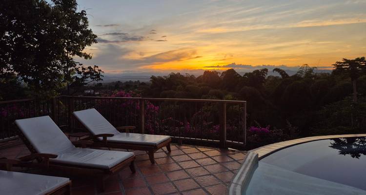 Two lounge chairs beside an infinity pool overlook lush forest and a colorful sunset horizon.