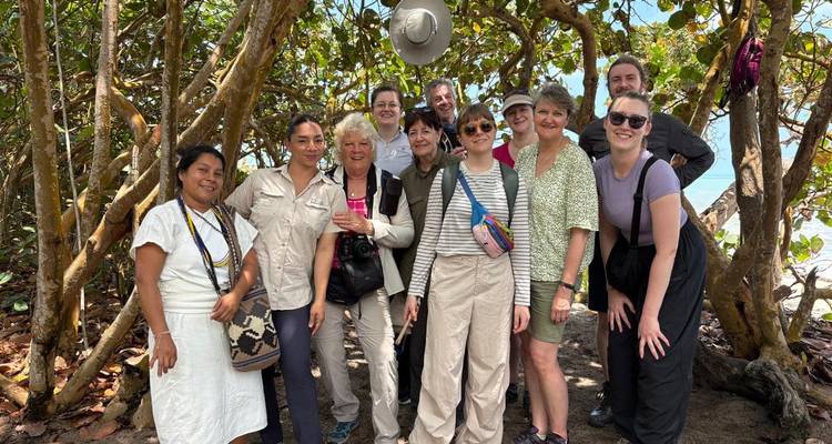Smiling tour group poses beneath tangled coastal trees with glimpses of turquoise water behind.