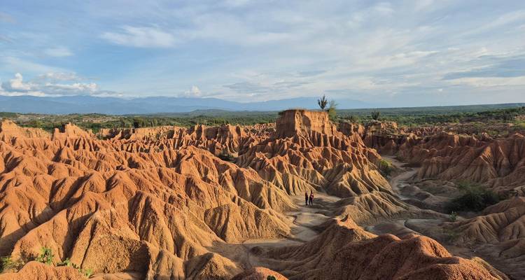 Vast reddish desert badlands with rugged ridges and two tiny visitors walking among the formations under a blue sky