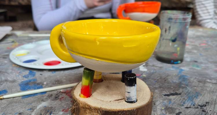 Bright yellow hand-painted ceramic cup being decorated on a messy art table with paint palette and brushes