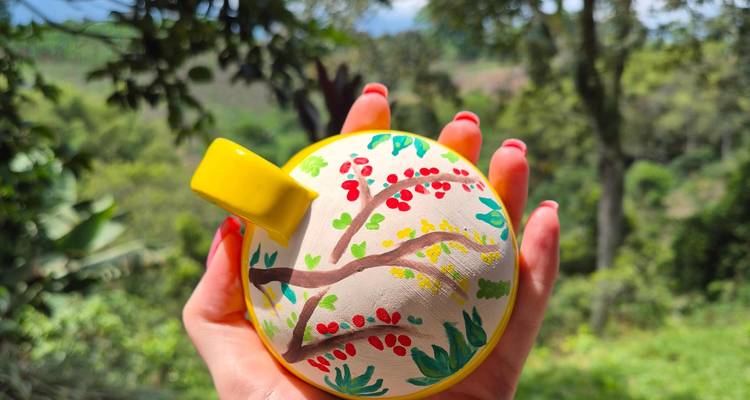 Hand with red nails holding a small, freshly painted ceramic pot against a lush green forest backdrop