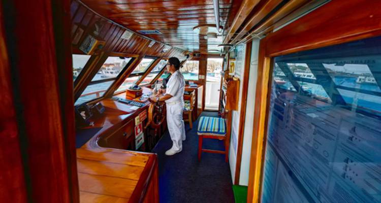 Scheepsbrug interieur met een bemanningslid aan het roer en ramen met uitzicht op de haven.