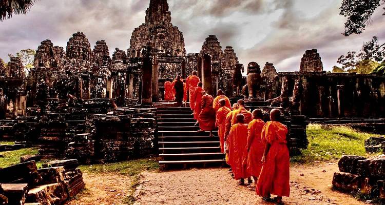 A procession of saffron-robed Buddhist monks ascends stone steps into the ancient Angkor temple complex under moody skies.