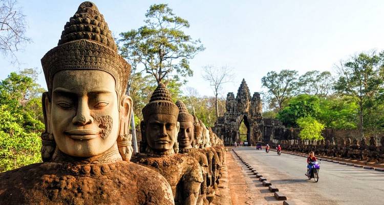 Massive stone guardian statues line the causeway leading to an ancient Khmer gateway beside a quiet road with motorbikes.
