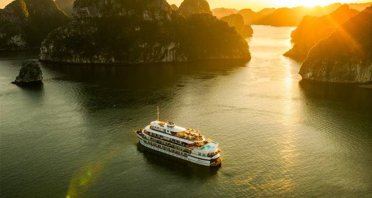 A cruise boat sails through emerald waters amid dramatic limestone karsts bathed in warm sunset light at Ha Long Bay.