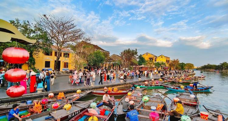 Crowds walk along Hoi An's riverside lined with colorful boats and lantern-adorned yellow buildings under a blue sky.