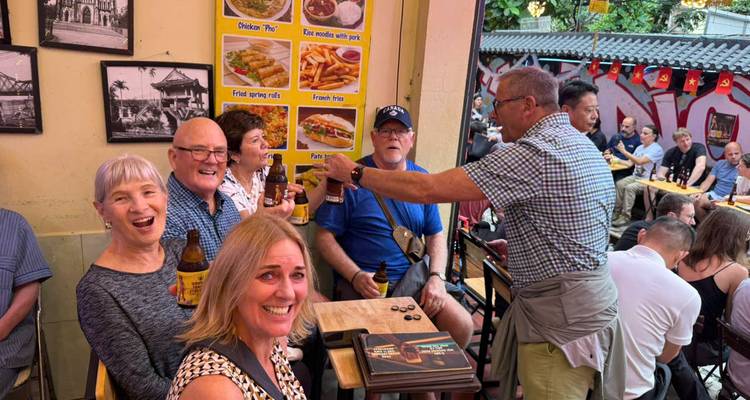 A cheerful group of older travelers enjoy beers at a bustling street-side eatery decorated with food photos.