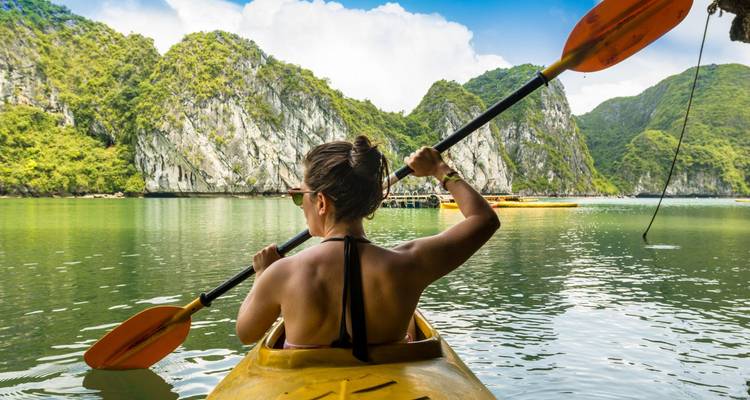 Una mujer rema en un kayak amarillo a través de aguas esmeraldas tranquilas rodeada de islas de piedra caliza empinadas cubiertas de selva.