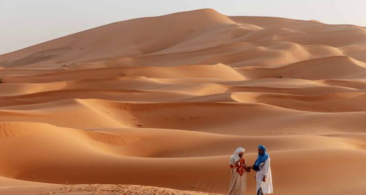 Eindeloze gouden zandduinen van de Sahara golven onder zacht avondlicht terwijl twee geroofde nomaden met elkaar spreken.
