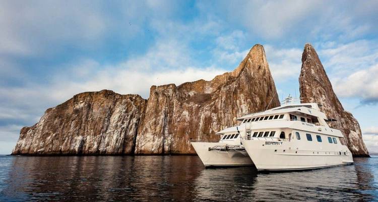 Catamaran blanco navegando frente a una imponente formación de roca volcánica que se eleva desde el mar.