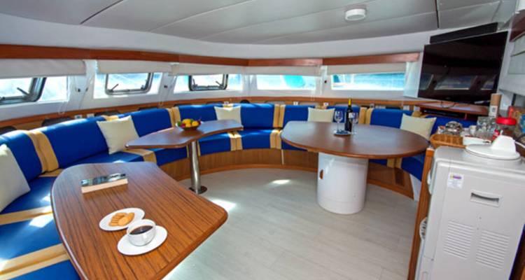 Salón moderno de yate con asientos azules curvados, mesas de madera, grandes ventanas y un televisor.