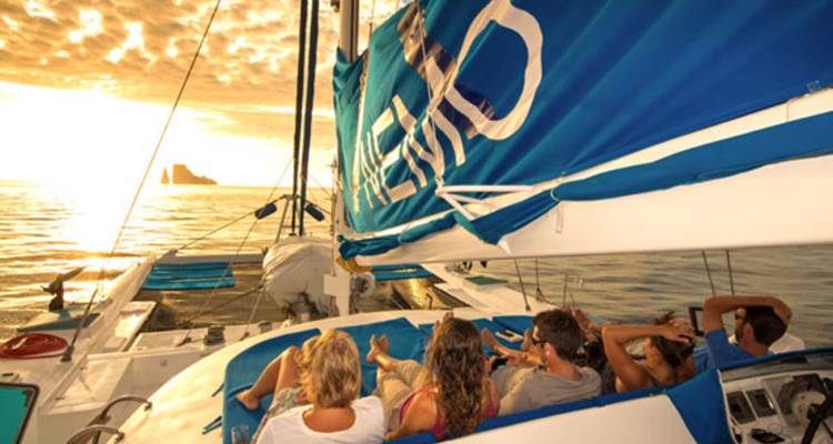 Un grupo de amigos relajándose en la cubierta de un catamarán mientras navegan hacia una puesta de sol de colores vibrantes.