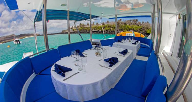 Configuración de comedor al aire libre con manteles blancos y cojines azules en un yate con vistas a aguas turquesas e isla verde.