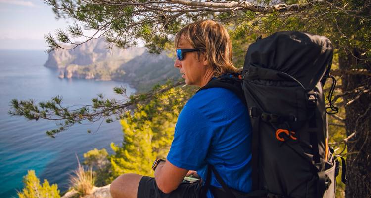Person with a backpack sitting on a rocky ledge overlooking the sea.