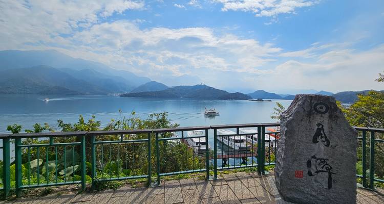 Vista tranquila del lago desde un mirador con barandilla con barcos turísticos y montañas brumosas en la distancia.